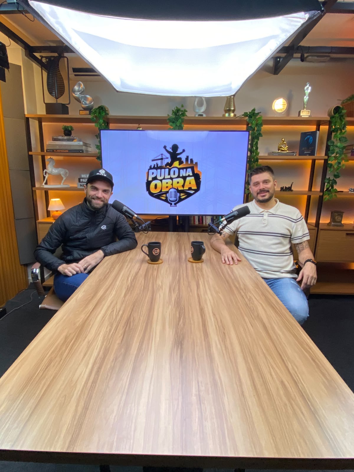 Os dois apresentadores do Pulo na Obra na mesa do estúdio, com a logo do programa na TV ao fundo